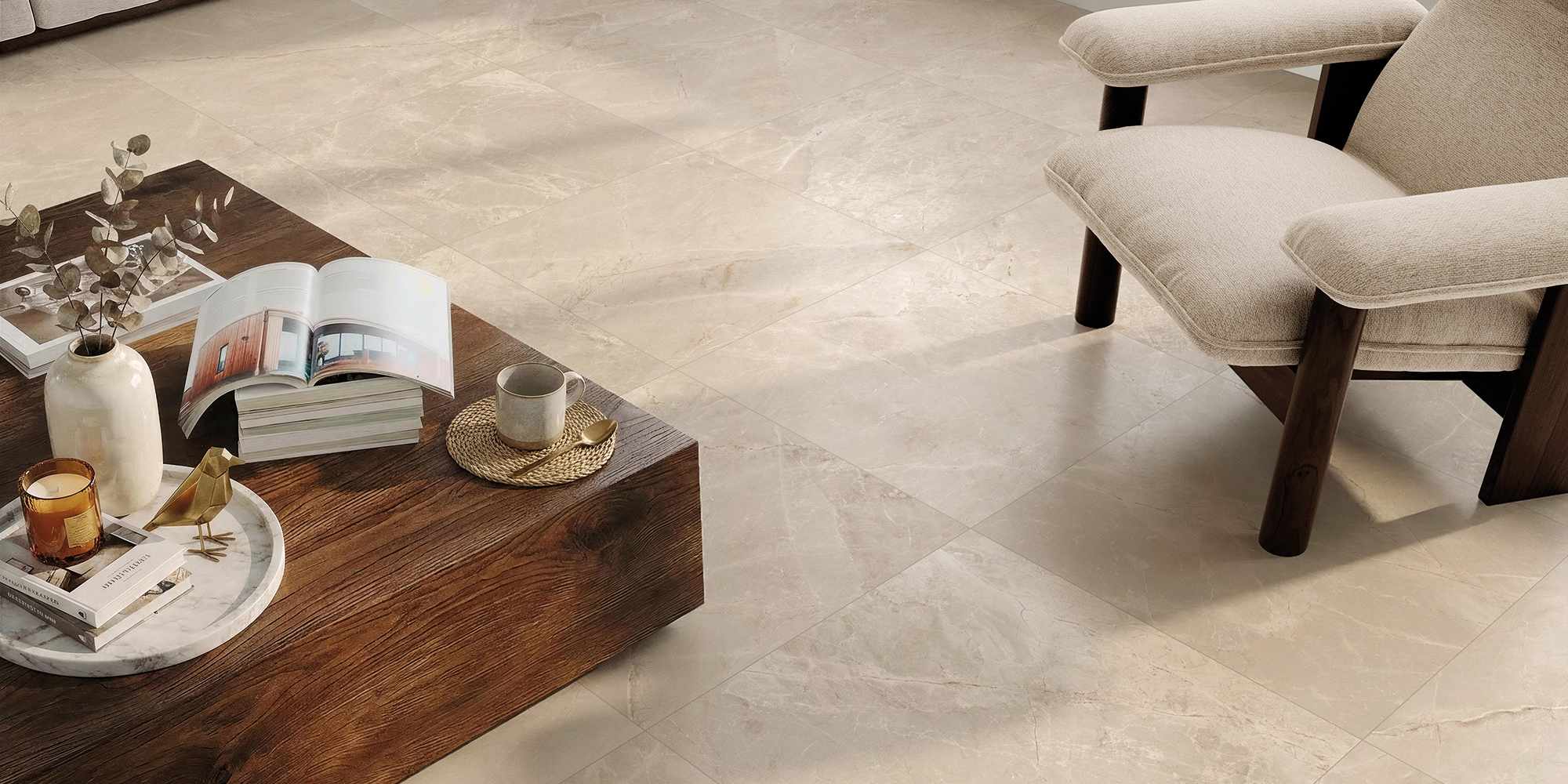 La collezione Amb Aurea, nella variante colore Elysium Sand in formato 60x60, valorizza il pavimento di un moderno e luminoso soggiorno attraverso lastre in gres porcellanato effetto pietra dalle calde sfumature beige. La posa ortogonale esalta le delicate venature naturali della superficie, su cui poggiano un tavolino basso in legno scuro finemente decorato e una raffinata poltrona in tessuto chiaro, creando un'atmosfera accogliente e di sobria eleganza.